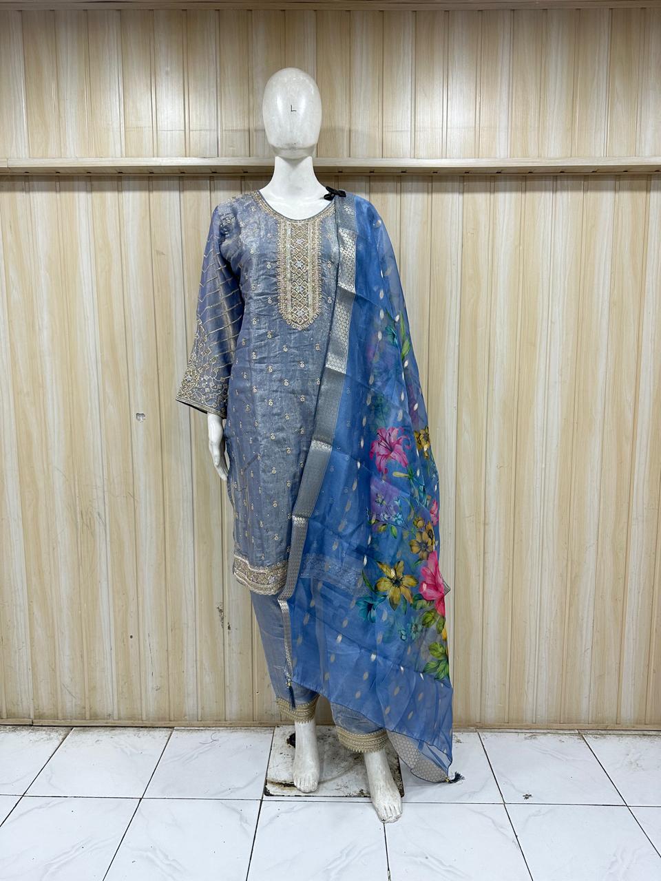 Mannequin wearing a blue embroidered outfit with a floral dupatta against a wooden panel background