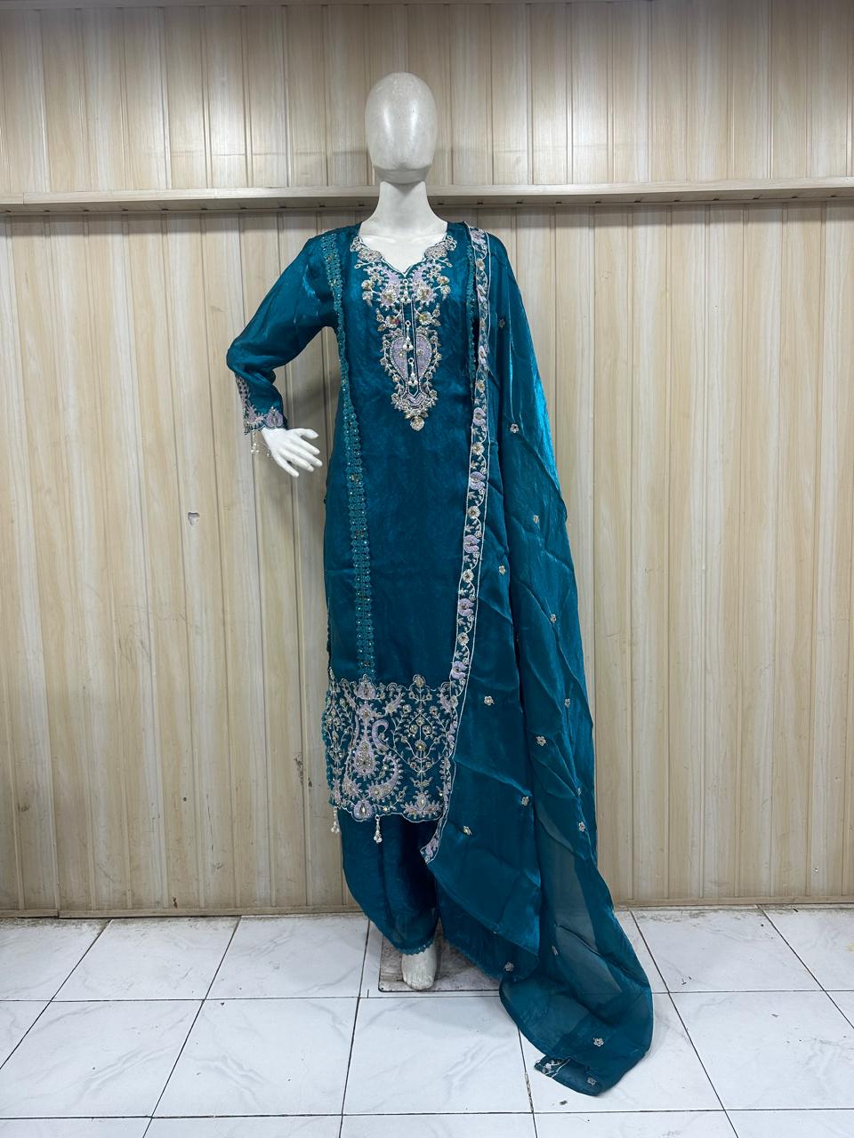 Teal traditional outfit with silver embroidery on a mannequin against a wooden panel background.