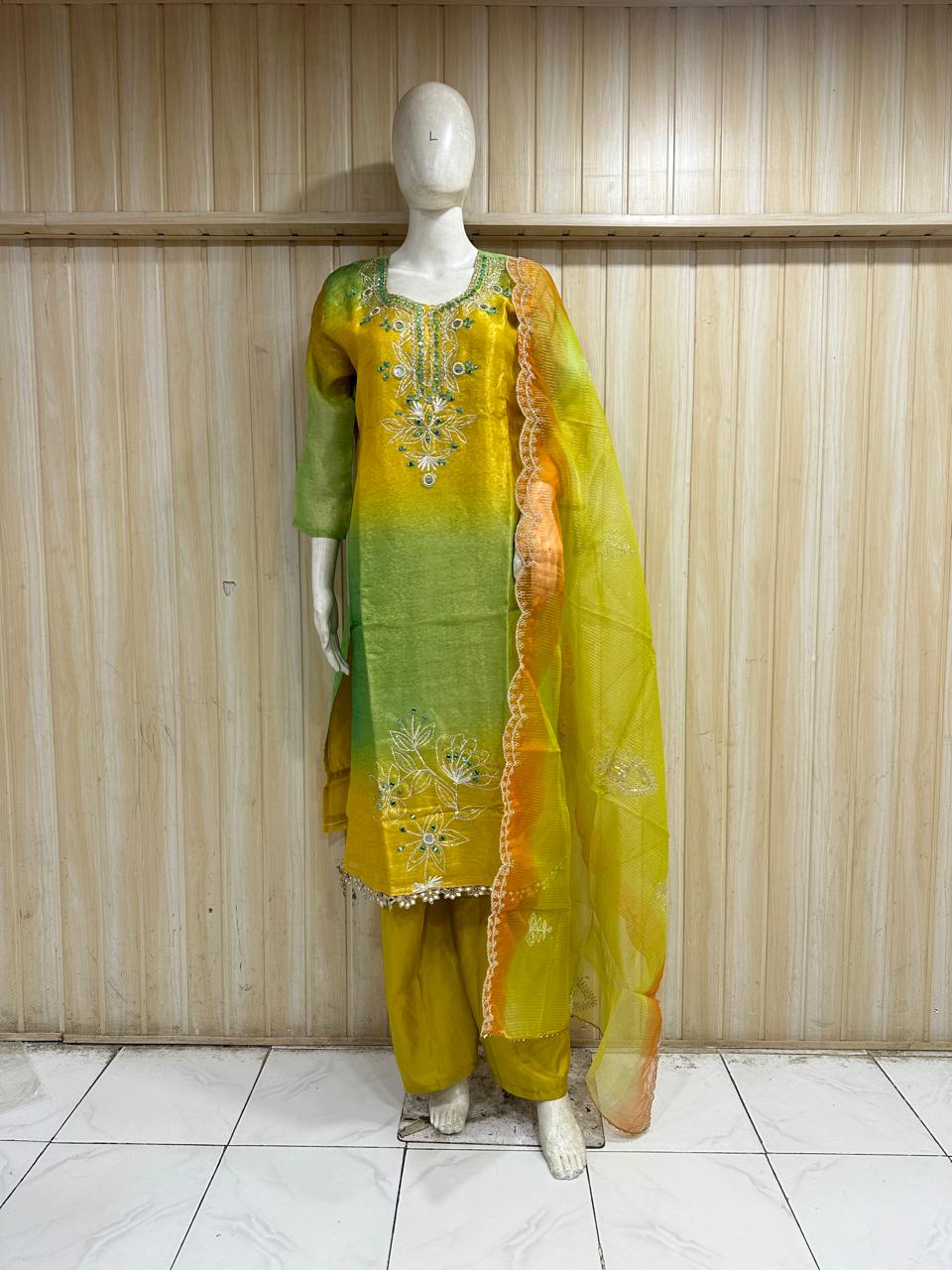 Mannequin wearing a yellow outfit with floral embroidery and a red floral dupatta against a wooden panel background.