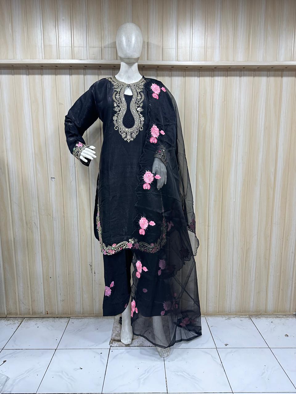 Black embroidered outfit with pink flowers on a mannequin against a wooden panel background.